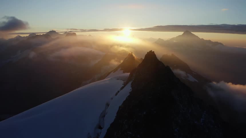 The sun is shining brightly on snow covered mountains with peaks high in a cloudy sky