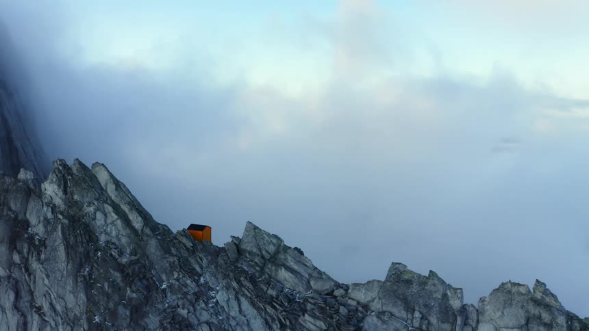 An aerial view of clouds covering a mountain range with a small orange building on the mountain