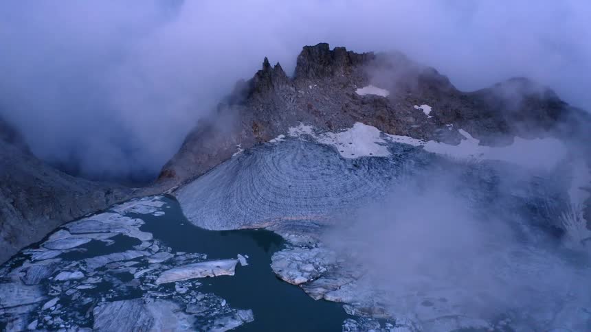 An aerial view of a pool of water and ice on a mountain with surrounding low clouds in the daytime