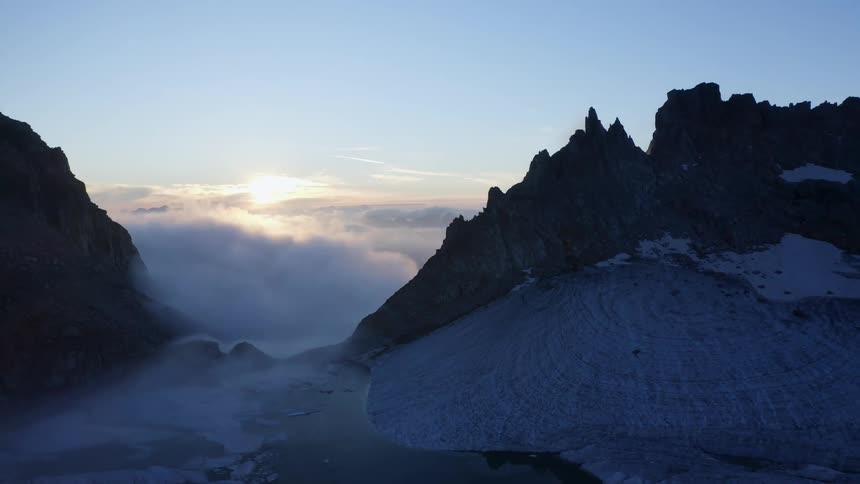 Sun shines above clouds through space occupied by water bordered by glacier and mountains