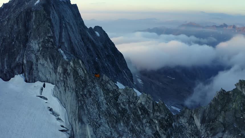 A small shelter sits on a narrow jagged mountain ridge with clouds and snow below