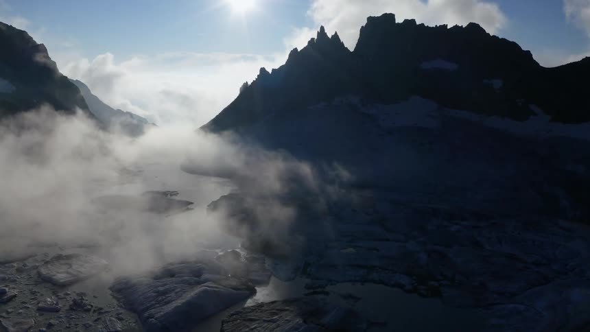 Fog floats over water dotted with ice and bordered by mountains on sunny day