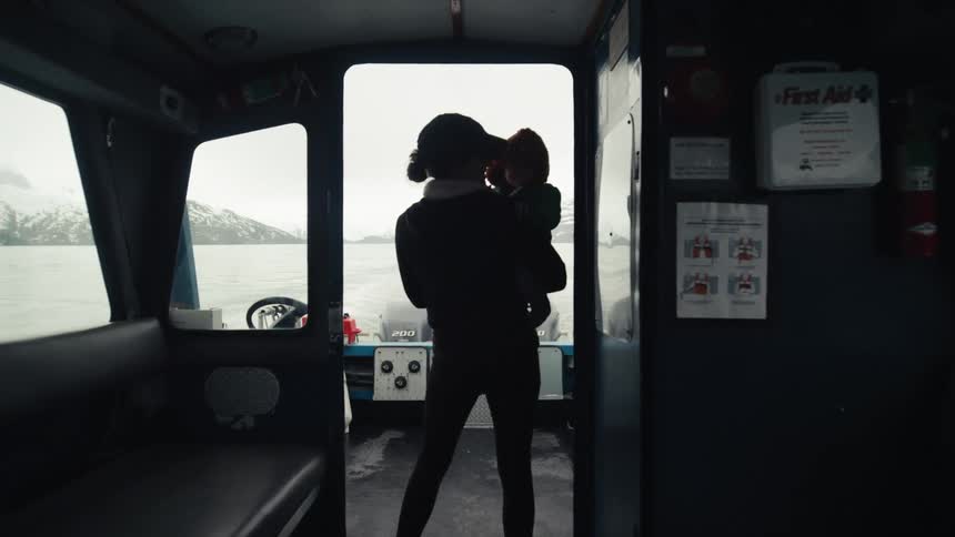 Mother stands outside boat cabin holding child with ocean and snowy mountains on horizon