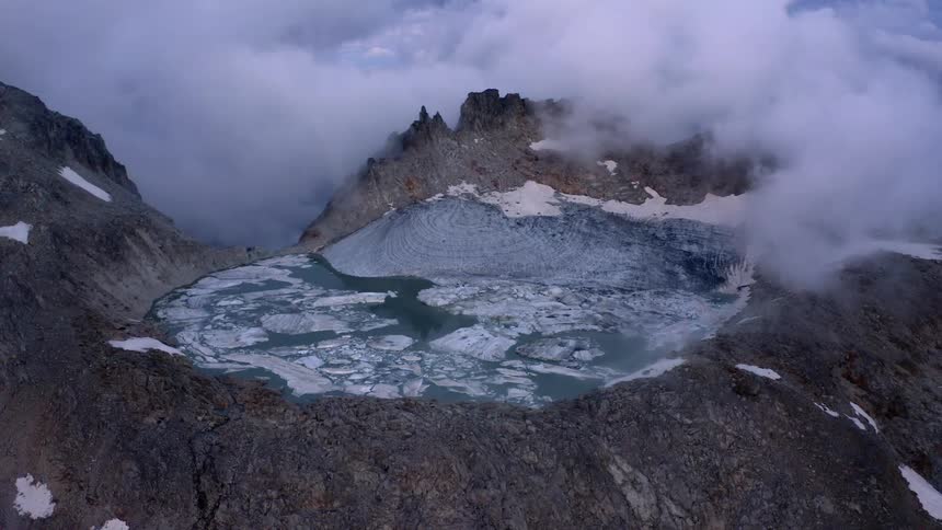 Ice floats on lake in crater under heavy fog bordered by snow dappled slopes