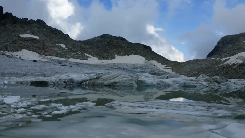 Ice covers shore and part of reflective water near mountains under cloudy sky