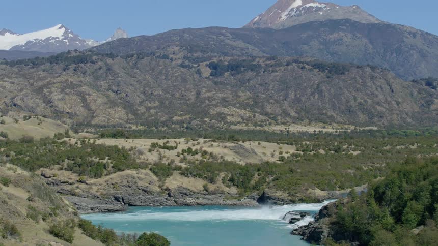 White water flows down short waterfall past forest with snow dappled mountains in background