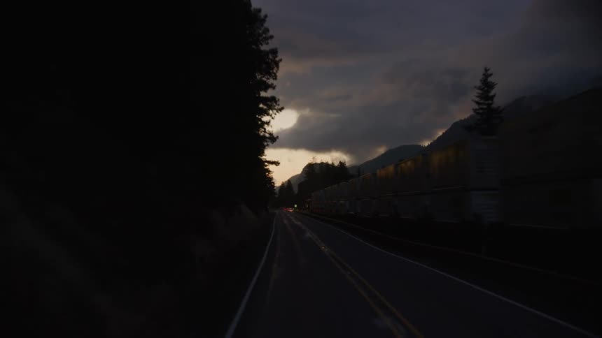 A narrow two lane highway with early morning sunrise clouds and a freight train as the car drives further and further along