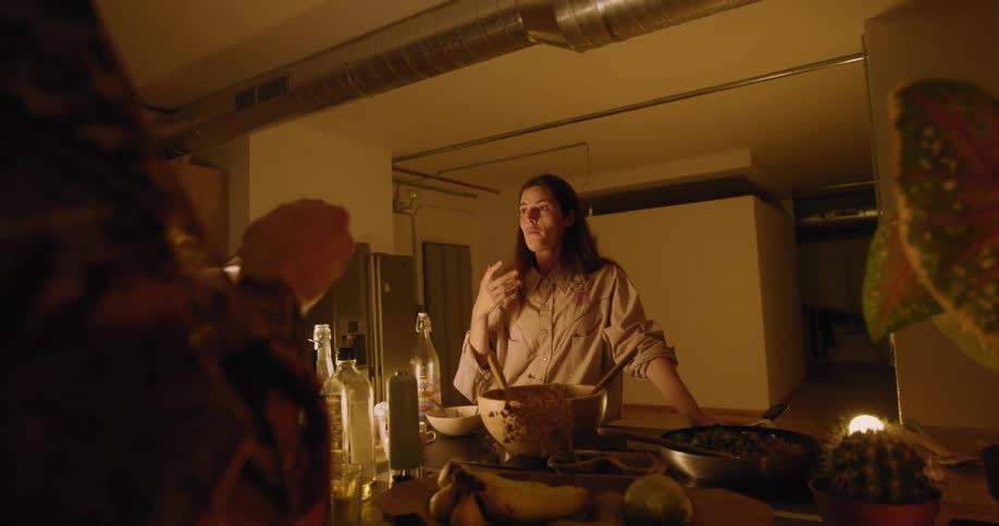 Woman stands in kitchen near stove talking to another woman eating from bowl