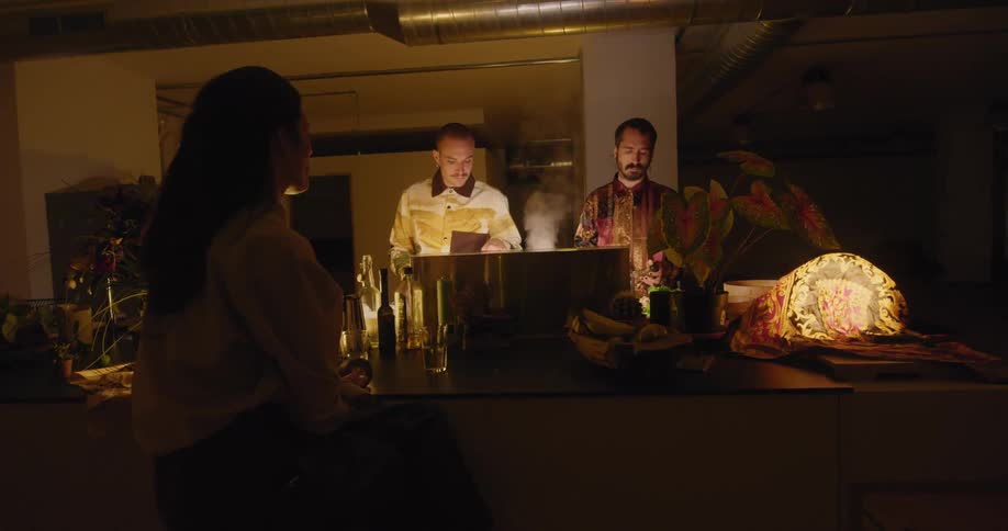 A woman is sitting at a bar while two men prepare food behind the bar at night