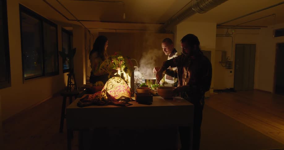 Two men are preparing food while a woman is sitting at a counter in a room at night