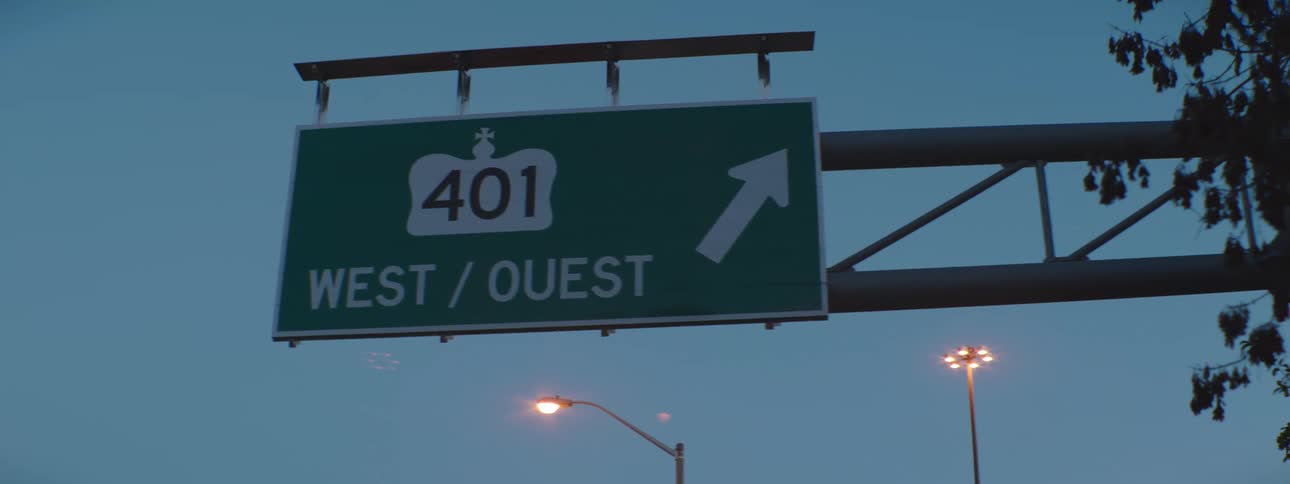 An exit sign for the 401 west with street lights glowing behind it