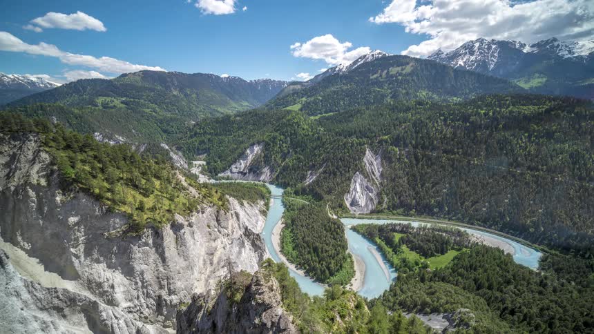 Clouds move across a bright blue sky above mountains and flowing stream