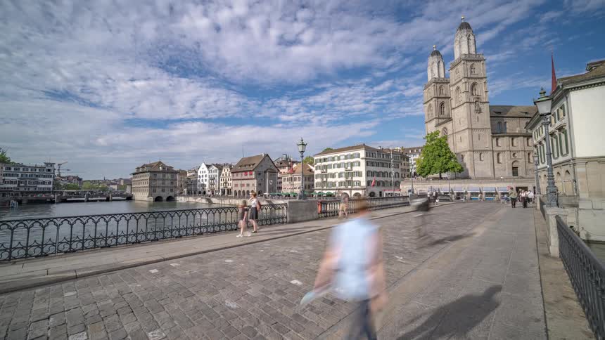 People and vehicles walk and drive on a bridge over a river near a historical city