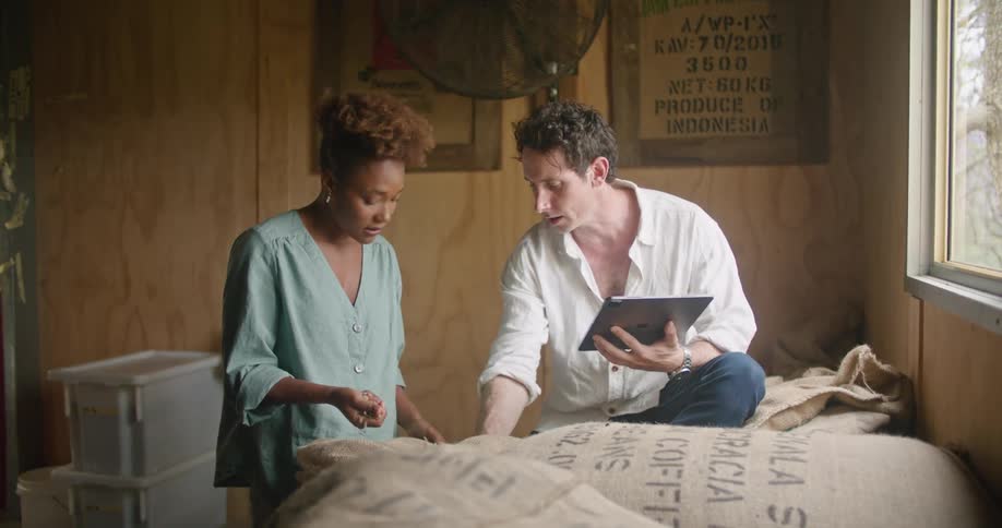 Woman with curly hairearring and light blue shirt checks beans next to man sitting on sacks using tablet in coffee warehouse
