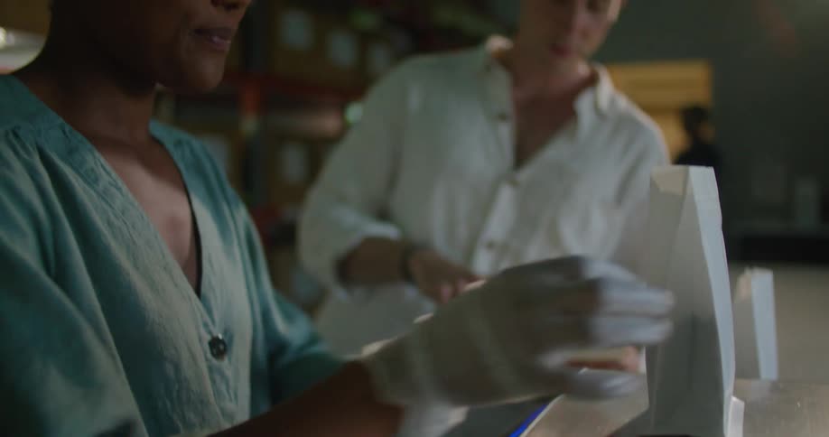 Woman with torquise shirt measures coffee beans next to man wearing white shirt and holding notebook in coffee warehouse