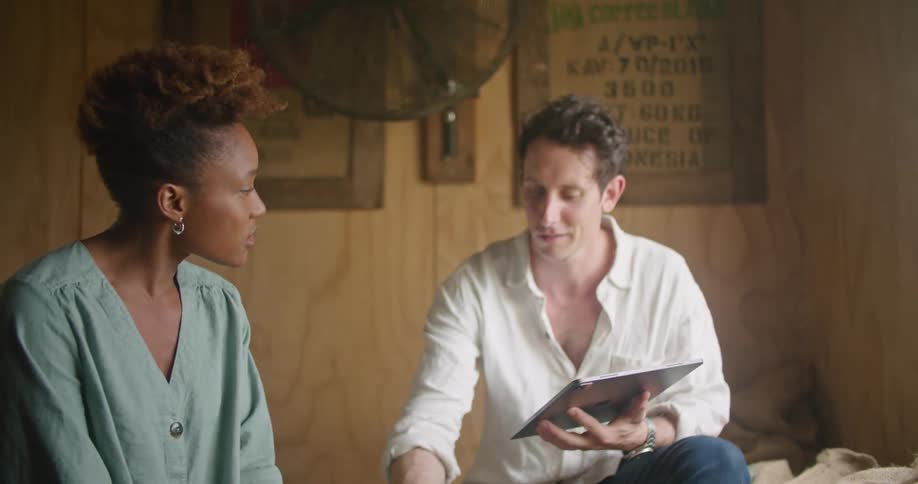Female worker with curly hair and and turquoise shirt and man wearing white shirt jeans and using tablet smell beans in warehouse
