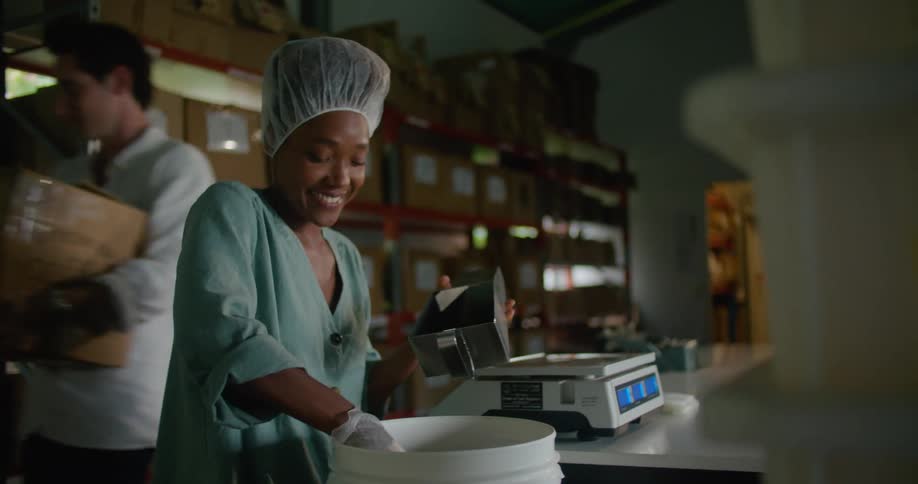 Woman with disposable hair bonnet gloves and turquoise shirt measures coffee beans while man carries box in coffee warehouse
