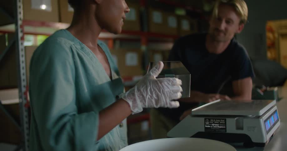 Female worker with curly hair and and turquoise shirt measures and packages coffee beans talking to blonde man with dark blue t shirt in coffee warehouse
