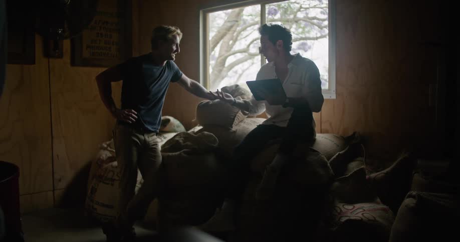 Man with white shirt and jeans sitting on coffee sacks works on tablet and talks with man with dark blue t shirt and brown pants carrying a sack of coffee in coffee warehouse
