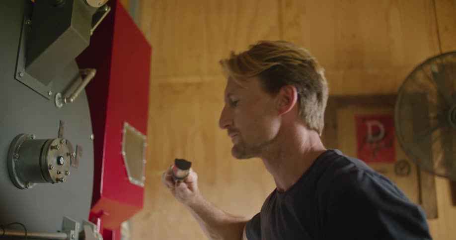 A man sticks a tool in to a large machine with coffee beans and pulls it out and smells it