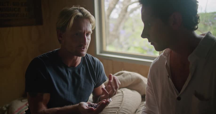 Two men sit on piles of burlap sacks as they discuss coffee beans