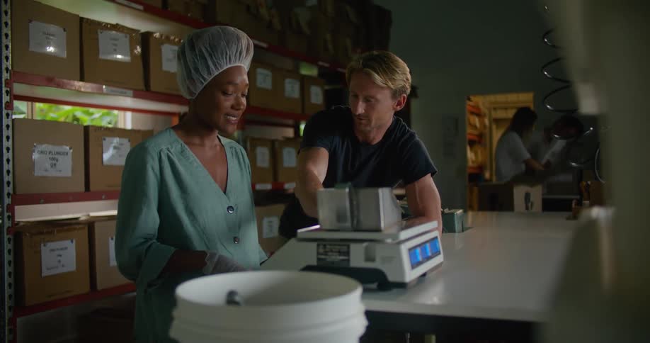 Woman with disposable hair bonnet gloves andturquoise shirt measures coffee beans while talking to blonde man wearing dark blue t shirt in coffee warehouse