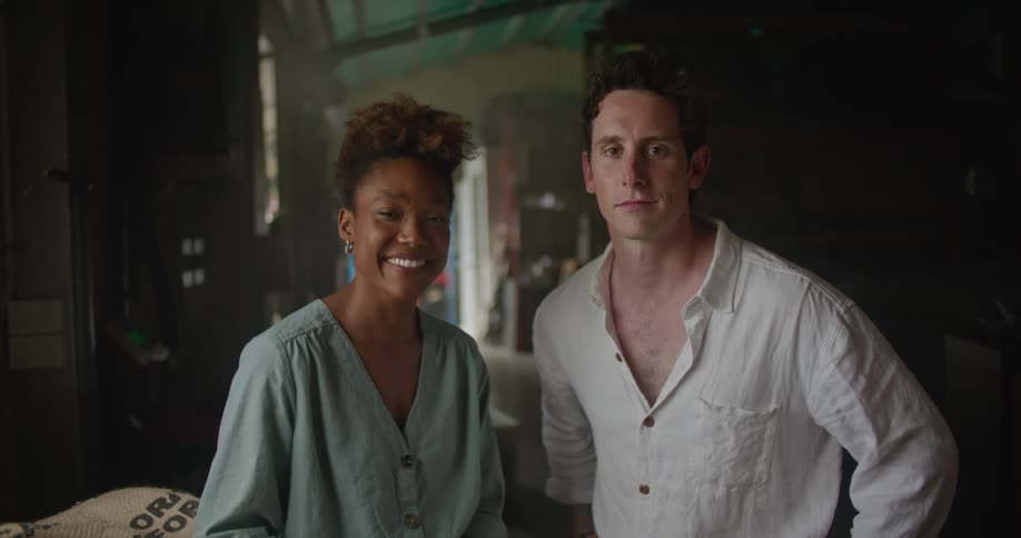 Man wearing white shirt and woman with curly hair and turquoise shirt stand next to coffee sacks and look at camera in coffee warehouse