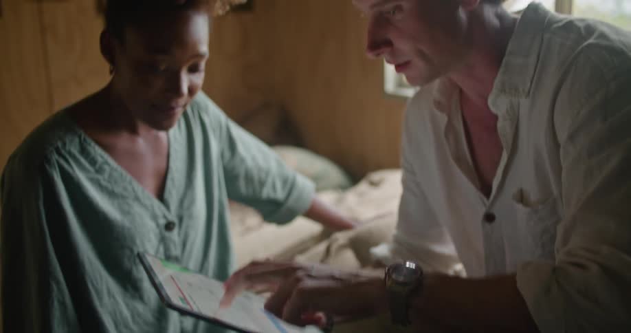 Man with white shirt and wristwatch sitting on coffee sacks works on tablet and talks to woman with curly hair and and turquoise shirt in coffee warehouse