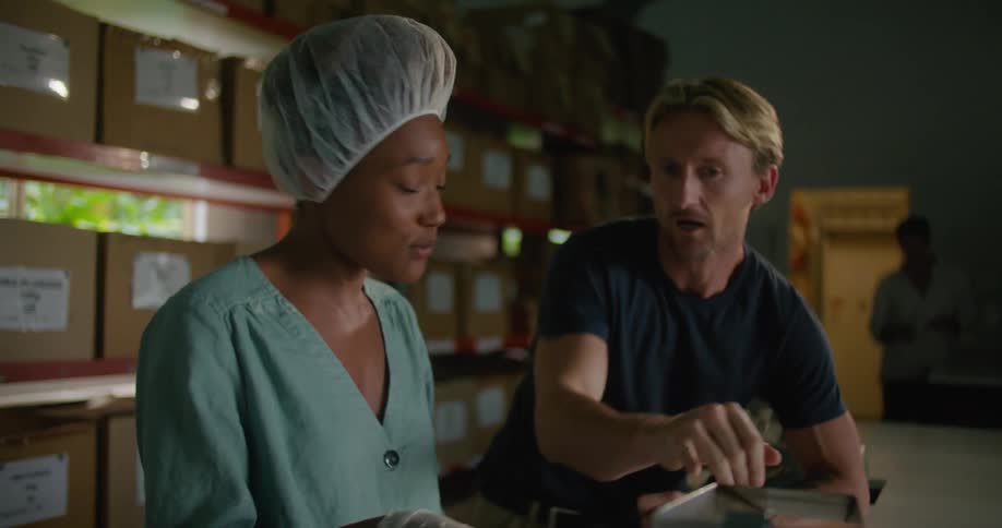 Female worker with hair bonnet and and turquoise shirt talks to blonde man wearing dark blue t shirt in front of shelves in coffee warehouse