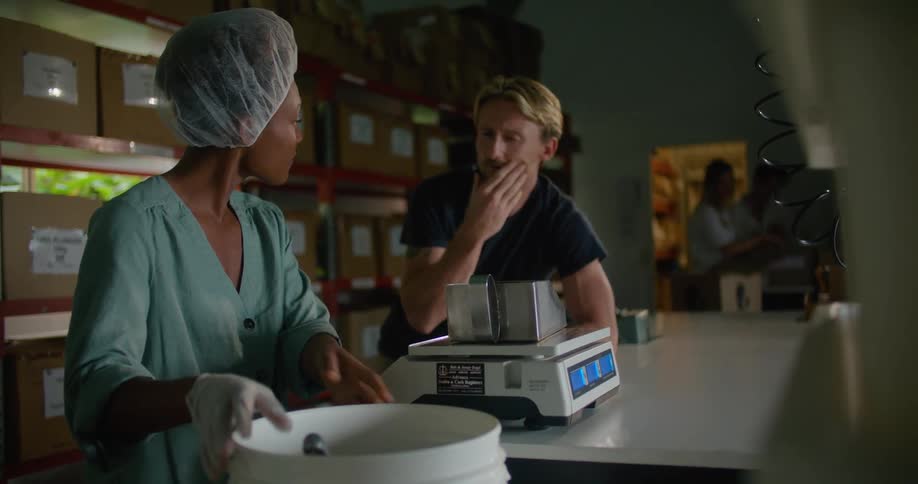 A man and woman in a cafe talk as the woman runs her gloved hand through a bucket of coffee beans
