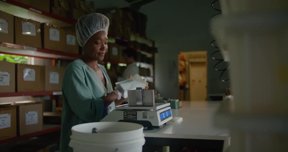 Woman with disposable hair bonnet gloves and turquoise shirt measures coffee beans while man carries box in coffee warehouse
