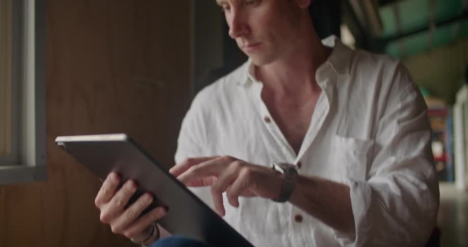 Man with white shirt and jeans sitting on coffee sacks and works on tablet in coffee warehouse