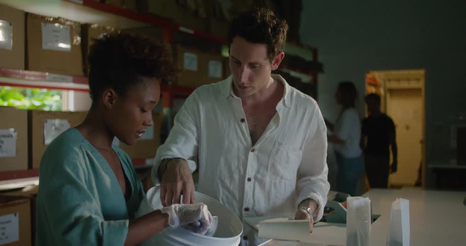Man with white shirt and brown wristwatch holds notebook and checks coffee beans in bucket talking to female worker with curly hair and turquoise shirt in front of shelves with boxes in coffee warehouse