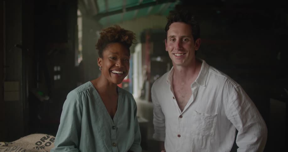 Woman with curly hair and and turquoise shirt and man wearing white shirt stand next to coffee sacks and look at camera smiling in coffee warehouse