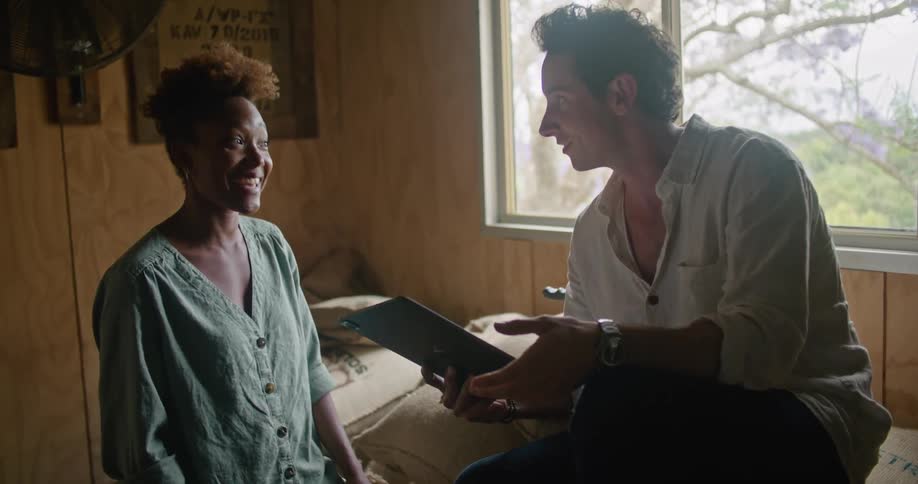 Female worker with curly hair and and turquoise shirt talks to man sitting on coffee sacks and working on tablet in coffee warehouse