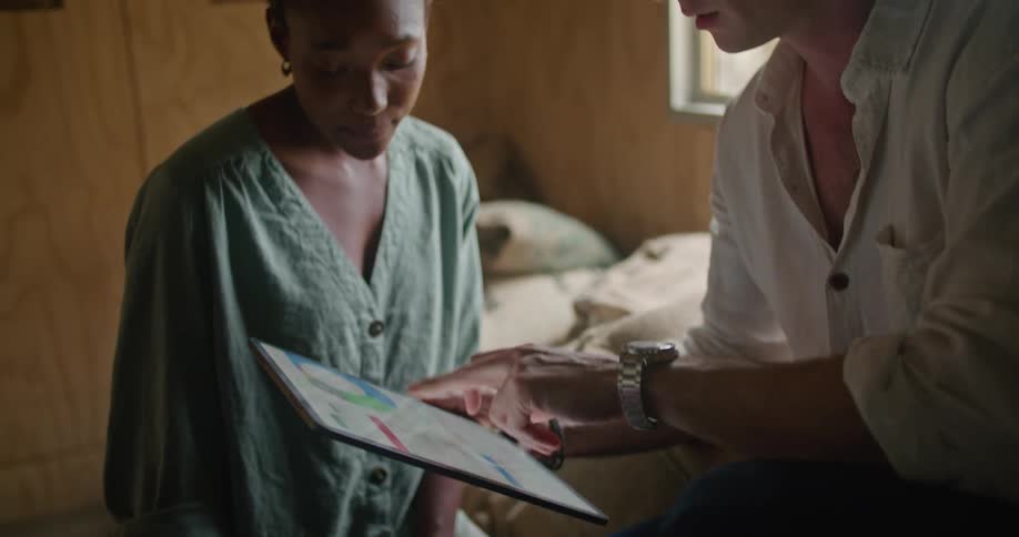 Man with white shirt and wristwatch sits on coffee sacks and works on tablet while talking to female worker with curly hair and and turquoise shirt in coffee warehouse