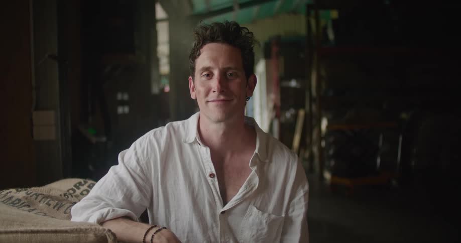 Man with white shirt leans against the coffee bags and looks at camera smiling in storage facility