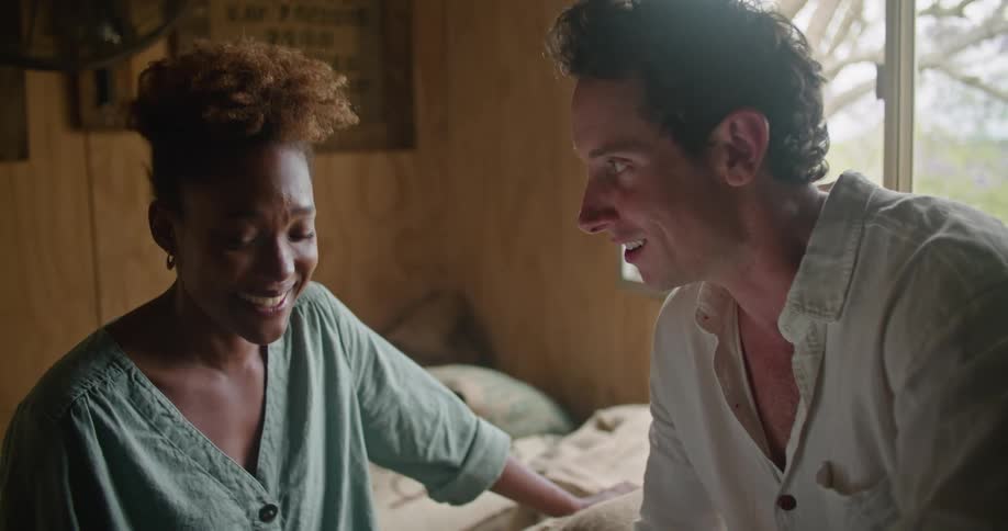 Man with white shirt and wristwatch sitting on coffee sacks works on tablet and talks with woman with curly hair and and turquoise shirt laughing in coffee warehouse