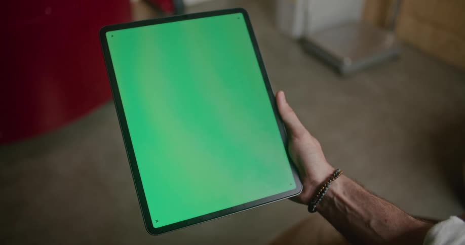 Man with white shirt and bracelet holds tablet with green screen in coffee warehouse