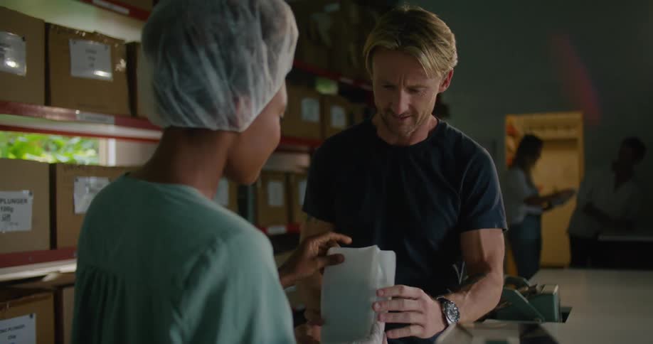 Blonde man smells coffee beans next to woman with hair bonnet and turquoise shirt in coffee warehouse