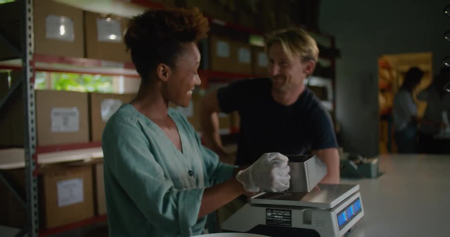 Female worker with curly hair and and turquoise shirt measures and packages coffee beans talking to blonde man with dark blue t shirt in coffee warehouse