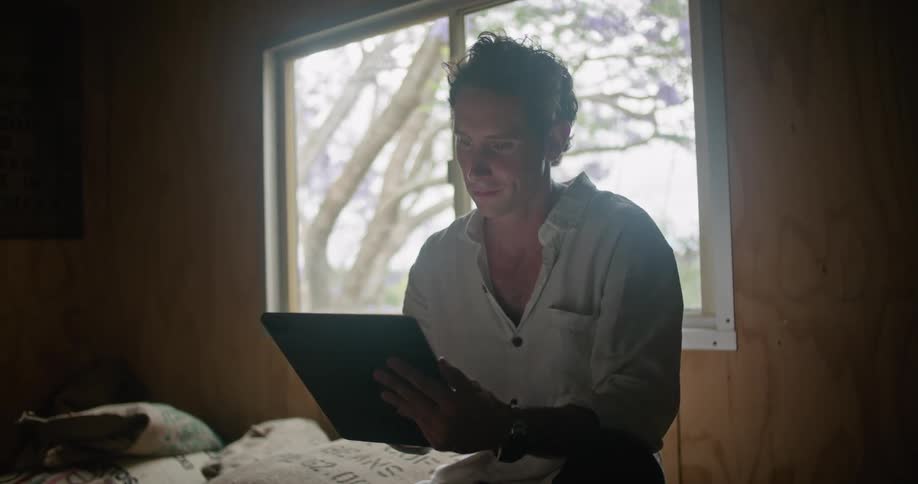 Man with white shirt sits on coffee sacks working on tablet in coffee warehouse
