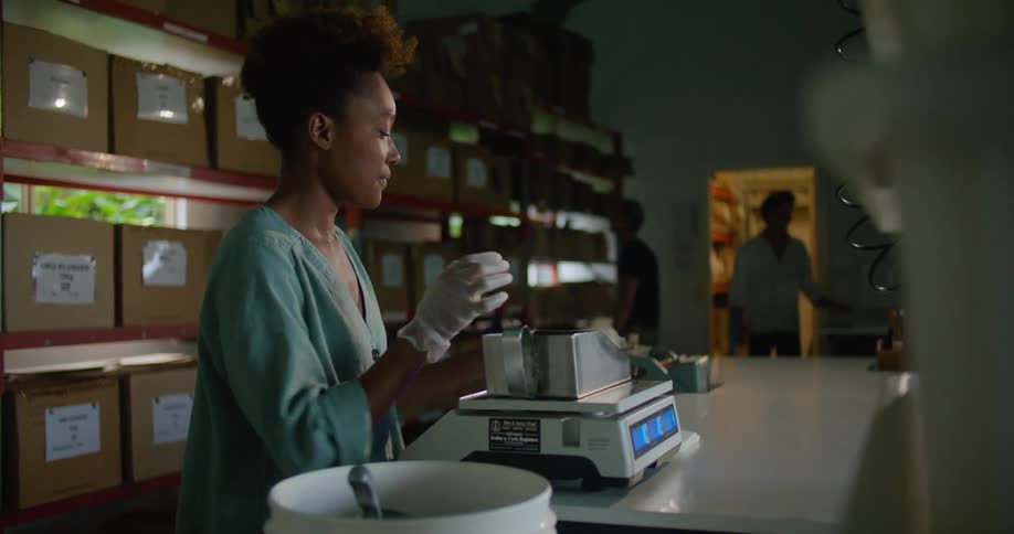 Female worker with curly hair and and turquoise shirt measures and packages coffee beans in front of the shelves with boxes in storage facility