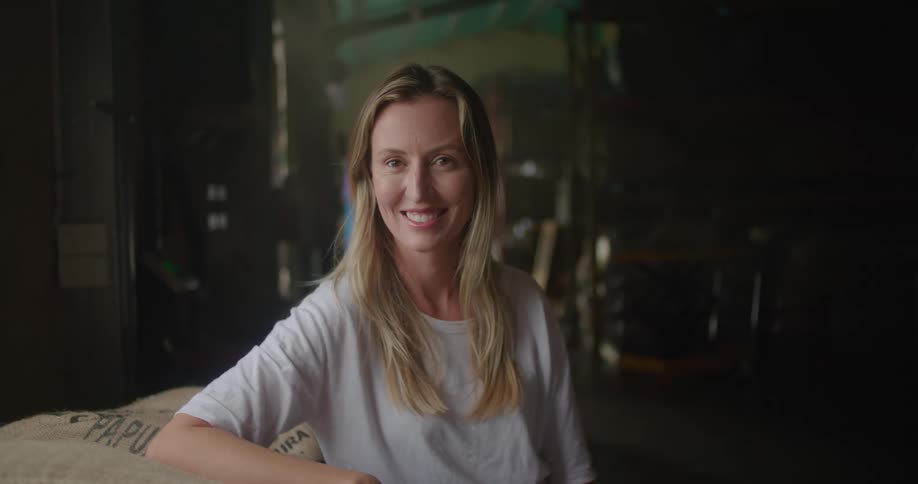 Blonde woman with white t shirt leans against coffee sacks and looks at camera smiling in coffee warehouse