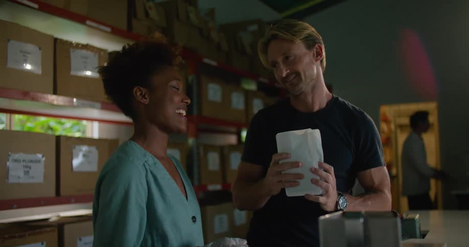 Female worker with curly hair and and turquoise shirt gives coffee beans to blonde man with dark blue t shirt to smell in coffee warehouse
