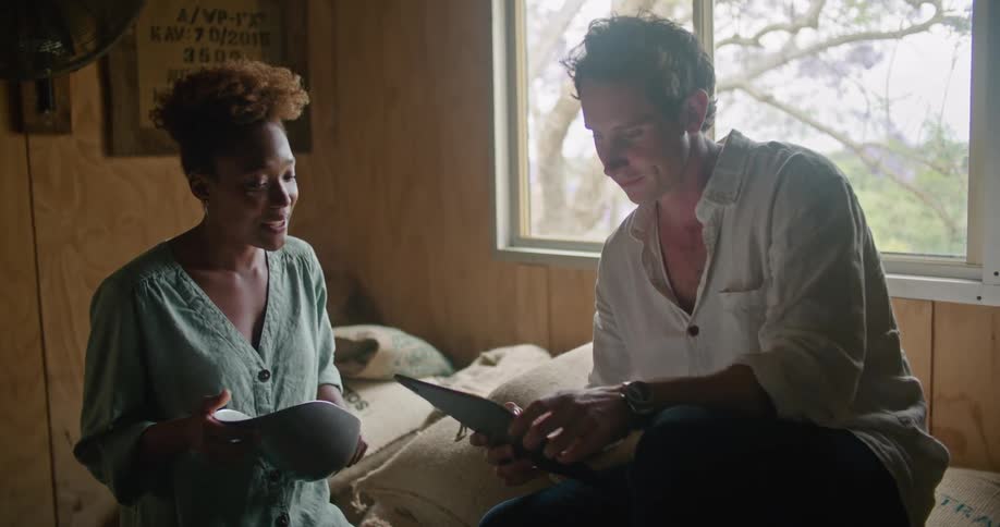 Man with white shirt sitting on coffee sacks works on laptop and talks to woman with brown curly hair andturquoise shirt in coffee warehouse