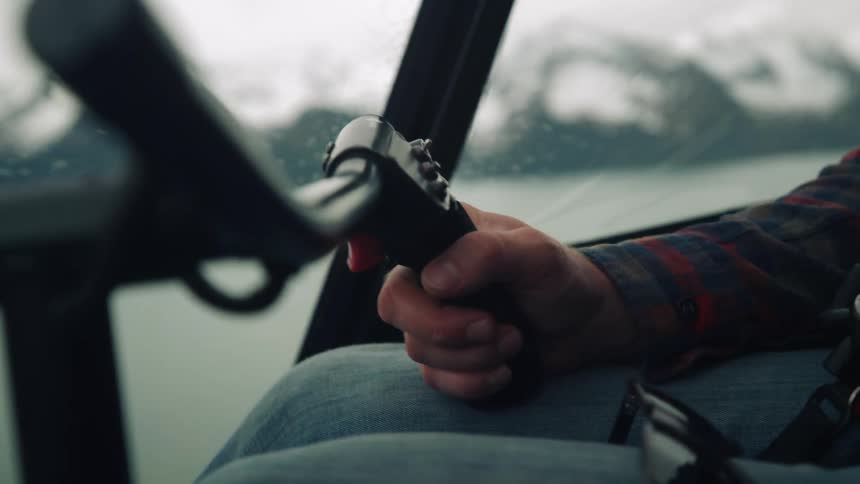 Male pilot with plaid shirt and jeans operates helicopter flying over water next to snowy mountains