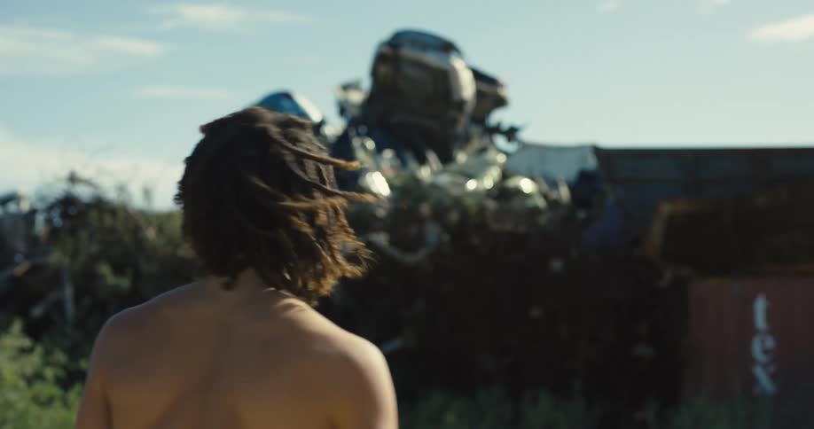 A shirtless boy walks over to a ridge looking out at a metal sculpture and tree tops