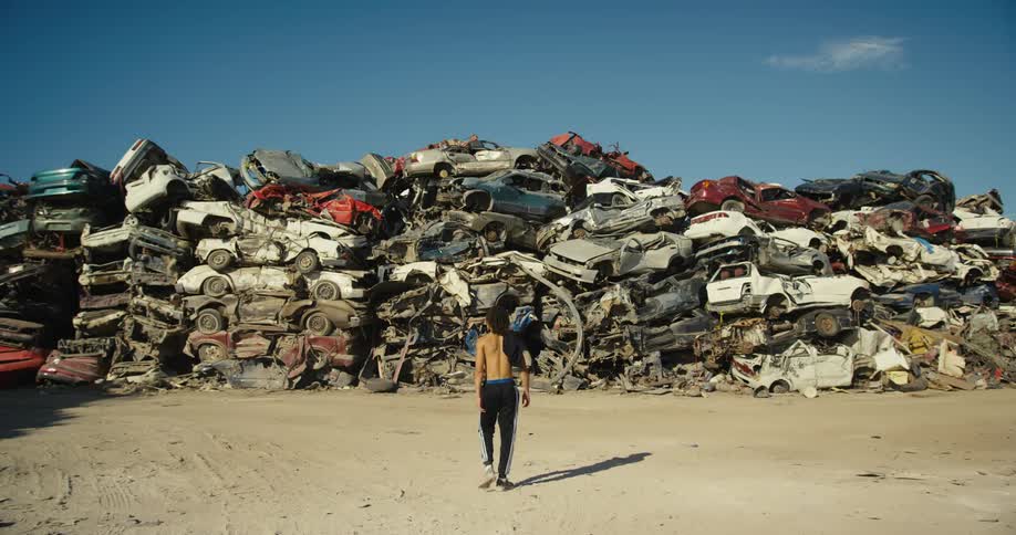 Topless youth walks across dirt lot to stand staring at hill of scraped cars