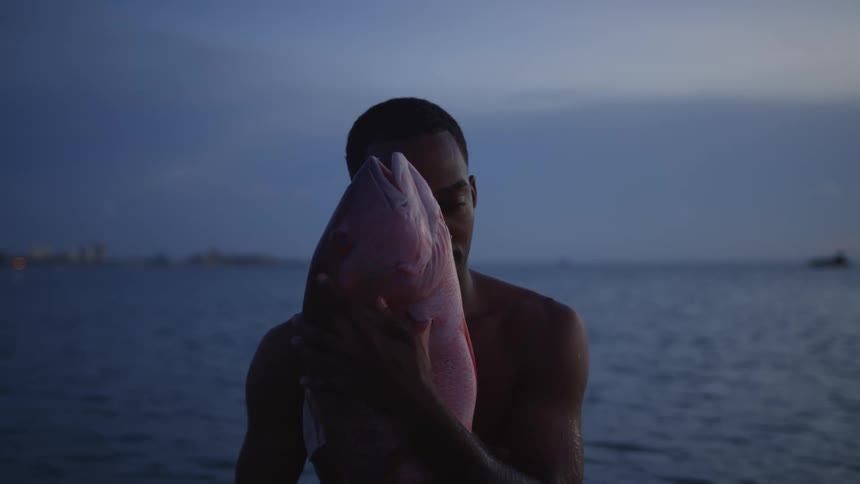 A young black man comes up out of the water holding a big fish in both hands